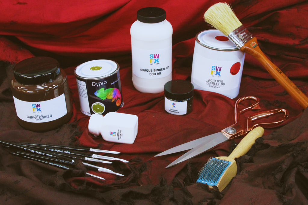 An assortment of dye tins, tools, paints and pigments arranged in front of a flowing red silk cloth